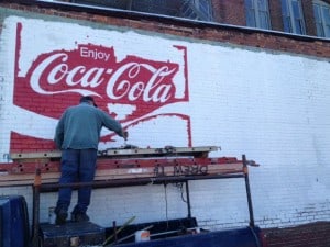 Signpainter applying red paint to coca cola sign on a white wall