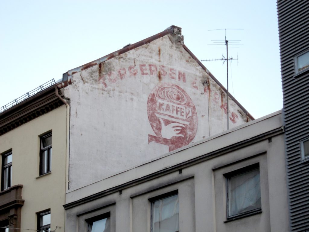 Fading painted sign with a business name arching across the top of the wall and a pictorial of a coffee cup underneath.