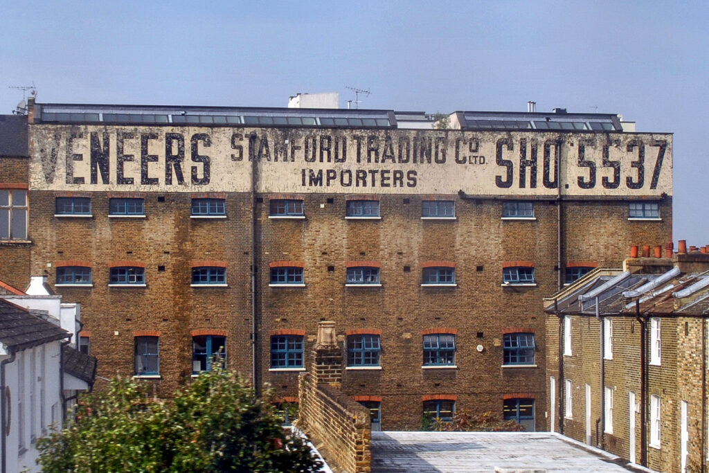 Large industrial building with a wide painted sign across the very top. The sign reads "Vaneers, Stamford Trading Co. Ltd., Importers, SHO. 5537" where the last bit is the old-format phone number for the business.