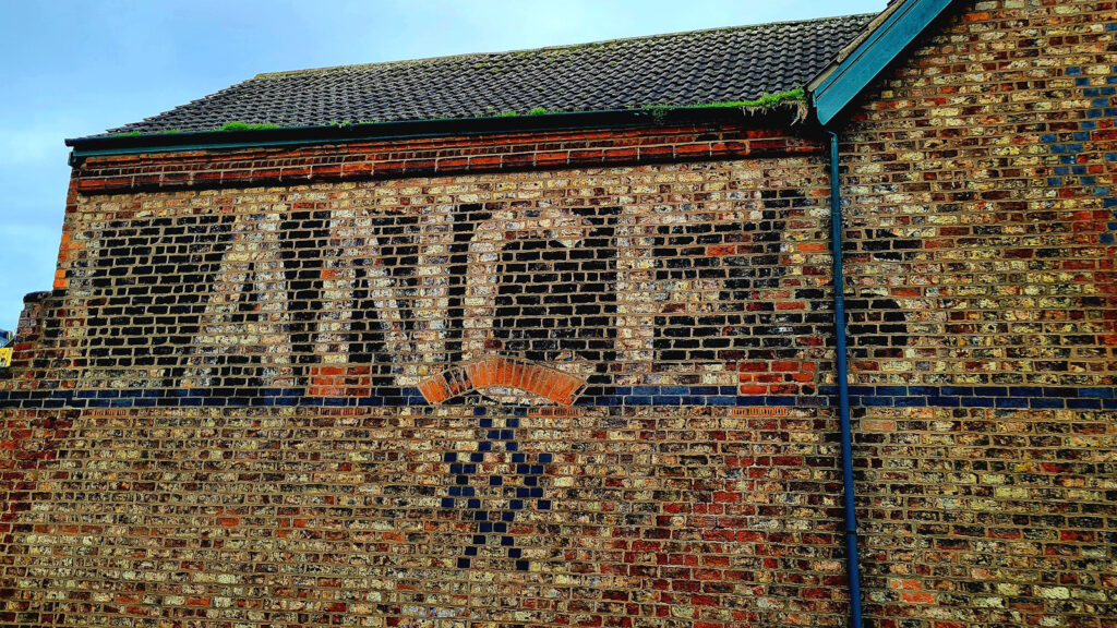 Fading remains of a painted sign on a brick wall. Only a partial fragment remains, which consistes of large white block letters set on a black background reading "Lances". A tiny part of the letter 'U' is visible to the left of this, with the wall beyond removed, along with the rest of the letters to complete the word "Ambulances".
