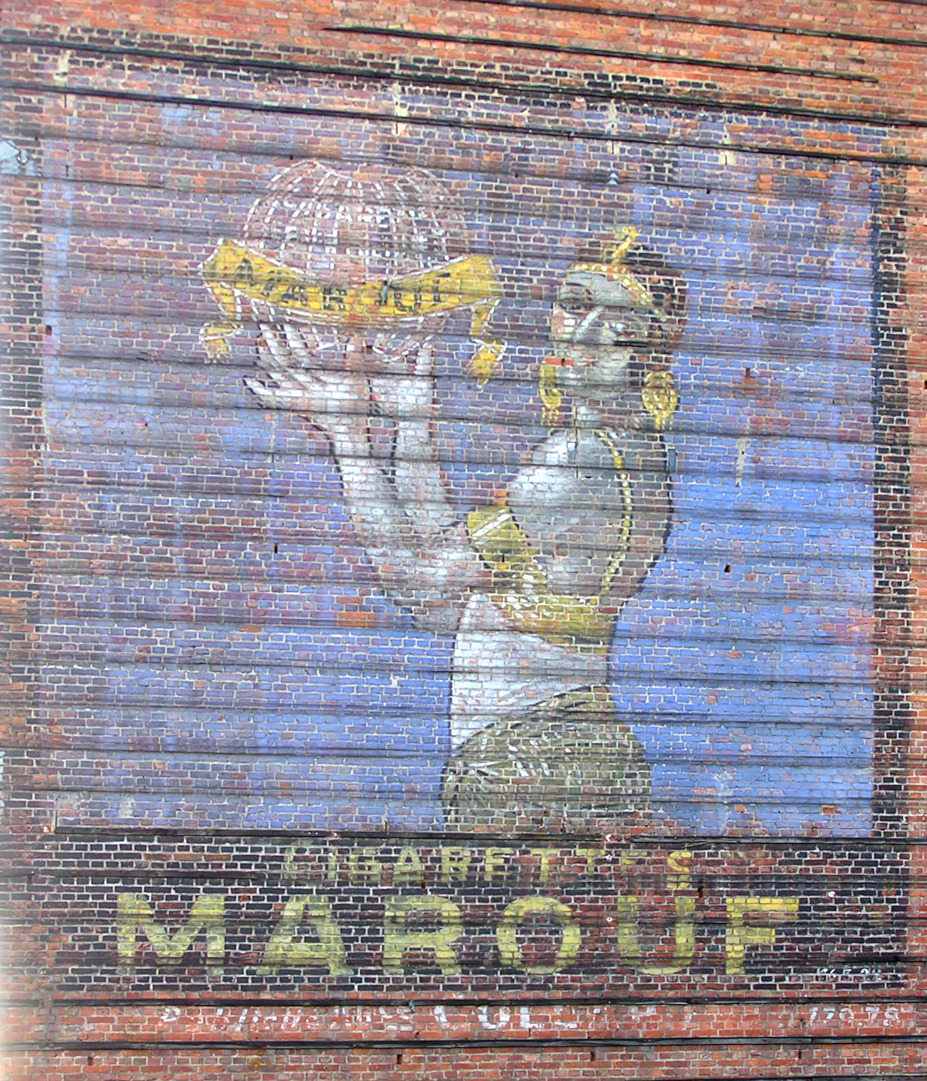 Fading painted advertisement on a red brick wall. It depicts a slender woman against a plain, bright blue background. She is shown from just below hip height, in side view, with her head turned to the left. She raises, with bent arms, a red-blue striped ball, in front of which is a yellow banner that reads "Marouf".

Towards the bottom of the sign is an extension of the black border that surrounds the whole composition, and set within this in yellow block letters is "Cigarettes Marouf".

There are some white letters below this, although are faded beyond legibility.