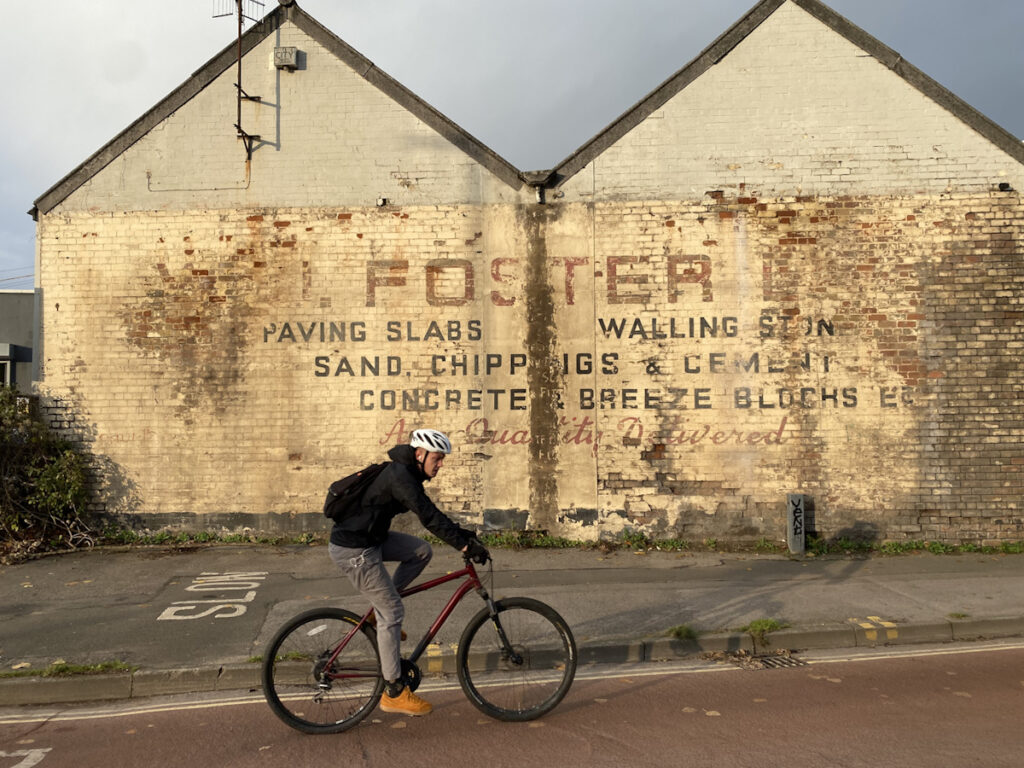 Twin gable end of a building with fading lettering on the wall advertising a builders merchants called Foster. In the foreground is a cyclist passing the wall unaware of being captured in the photograph.