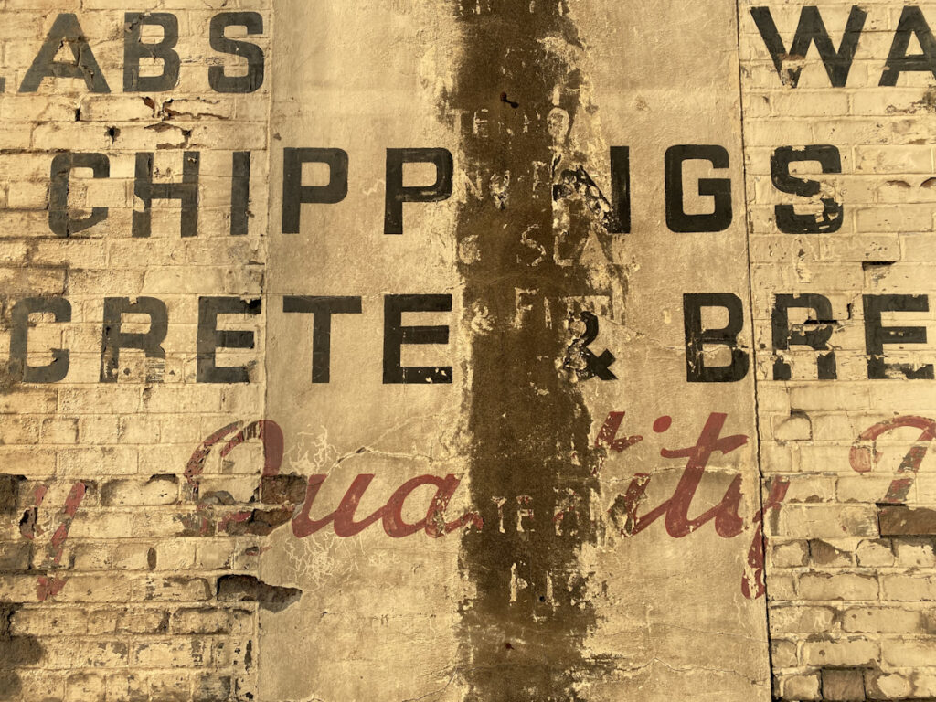 Close-up photo of a portion of a wall with painted black and red lettering fading on a cream background. Running vertically down the middle of the ghost sign is a strip that has decayed a lot more revealing fragments of smaller lettering beneath the main sign.