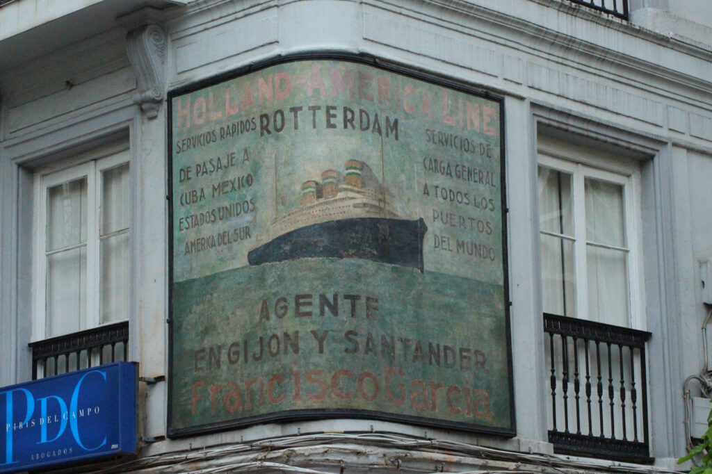 Touched up fading painted sign on a curved wall on the first floor of a corner building. It shows a large steamship sailing on the ocean, surrounded by lettering advertising the services of the Holland-America Line.