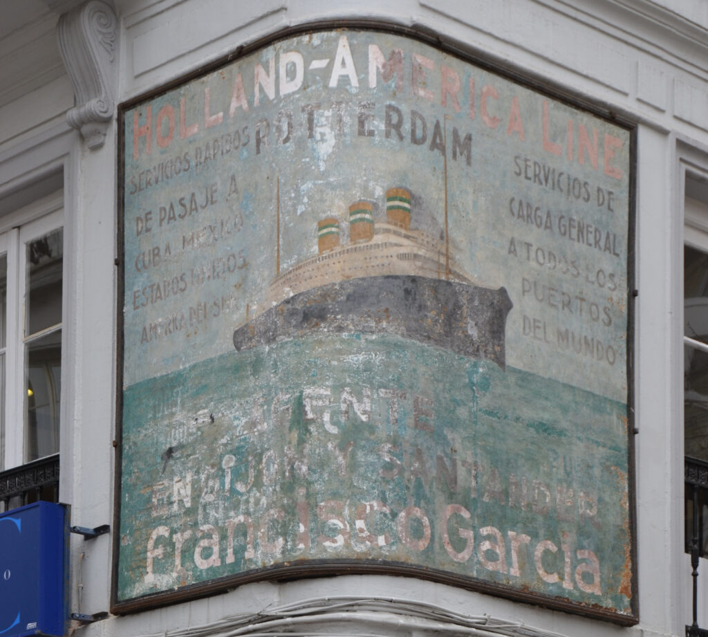 Fading painted sign on a curved wall on the first floor of a corner building. It shows a large steamship sailing on the ocean, surrounded by lettering advertising the services of the Holland-America Line.