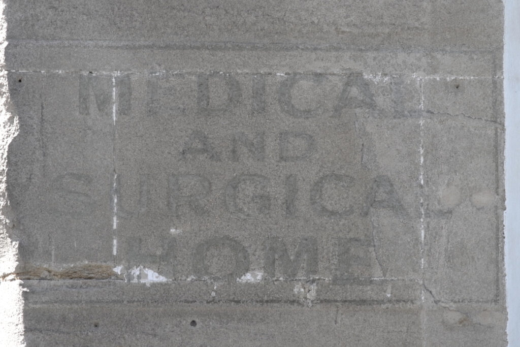 Fading painted letters on a slab of stone on a wall. They are painted in a simple block style, surrounded by a thin right-angled border, and read "Medical and Surgical Home".