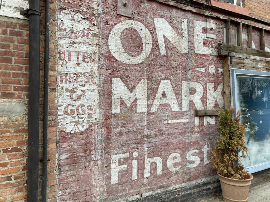 Brick wall with a fading painted advertisement on it, which isn't visible in its entirety. The sign, on a faded red background, consists of two main parts: a panel on the left that is red lettering out of a white panel reading "Finest Butter, Cheese, & Eggs"; and the main part of the sign which is partially obscured by a later window, but which once read "Oneida Market in Finest ____".