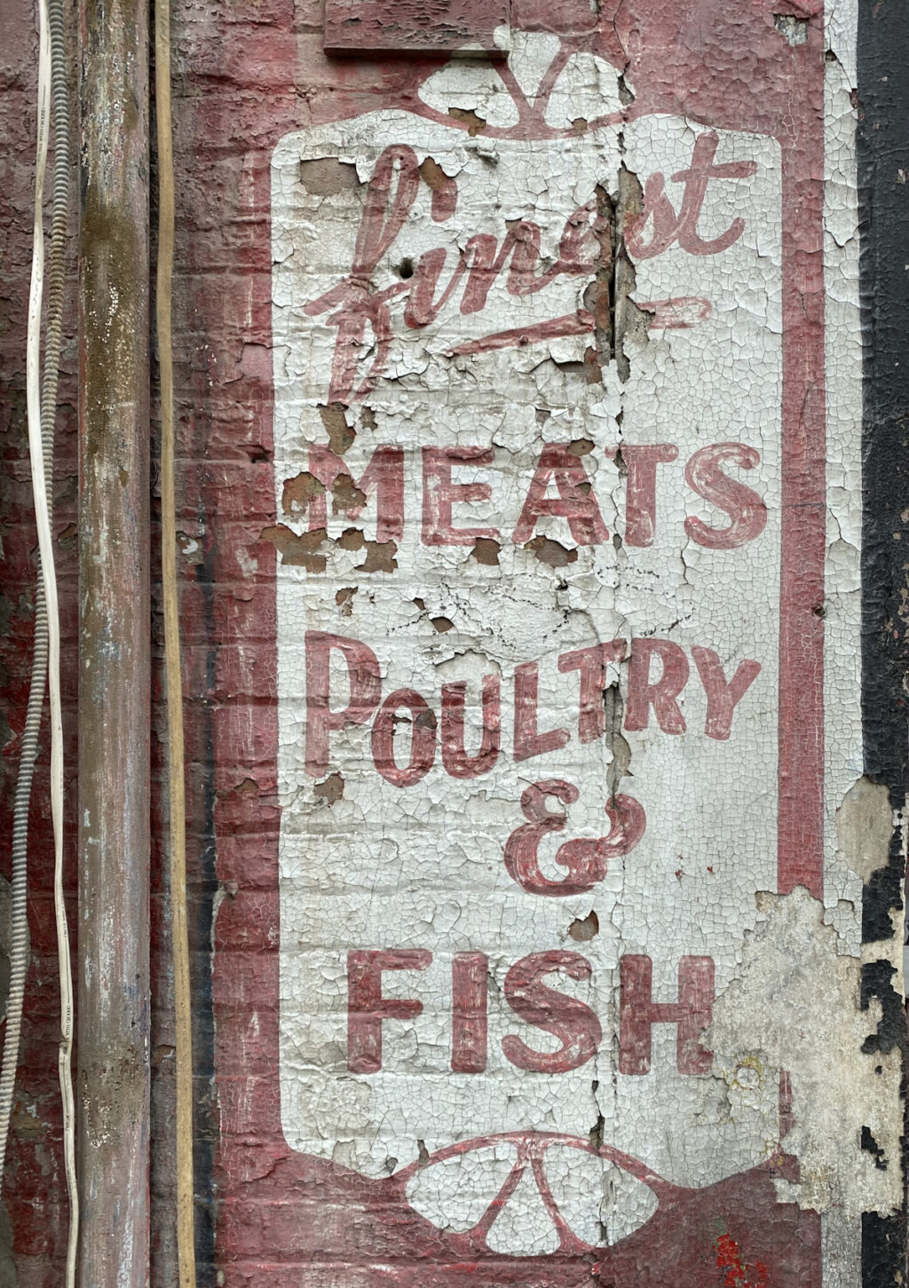 Fading painted sign on a brick wall. It is set within a white panel on a red background, with red lettering that reads: "Finest Meats, Poultry & Fish".
