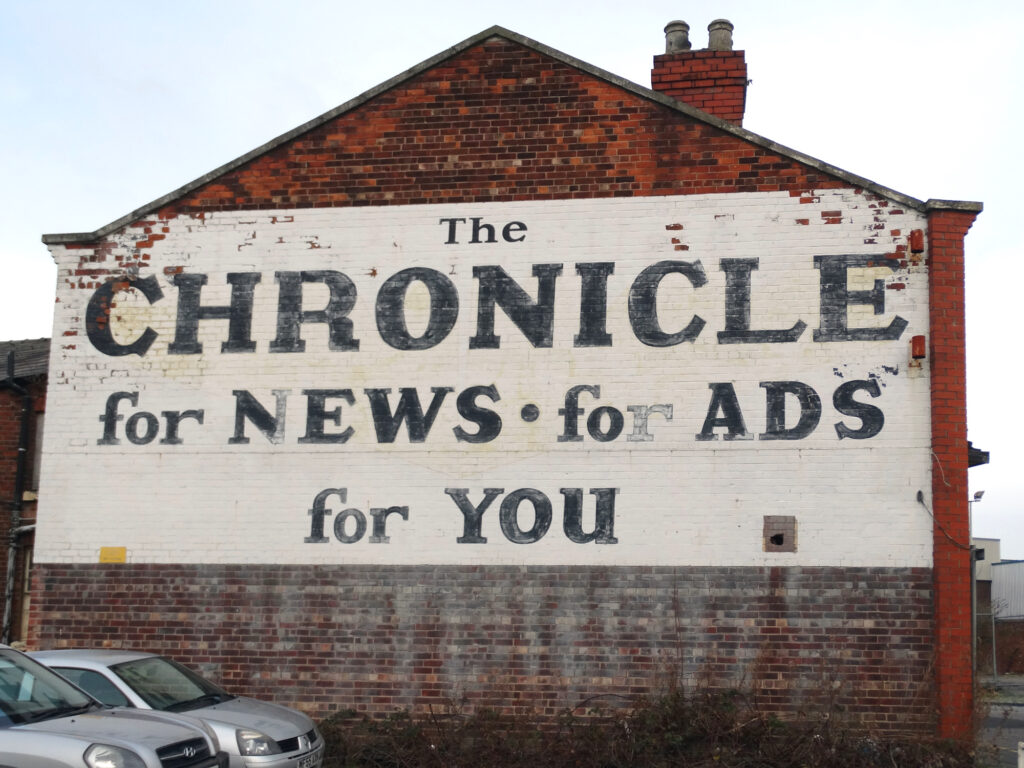 Painted sign on a brick gable end. It fills the full width of the wall, and about half its height, with an off-white background and black seriffed lettering that reads "The Chronicle, for News, for Ads, for You".