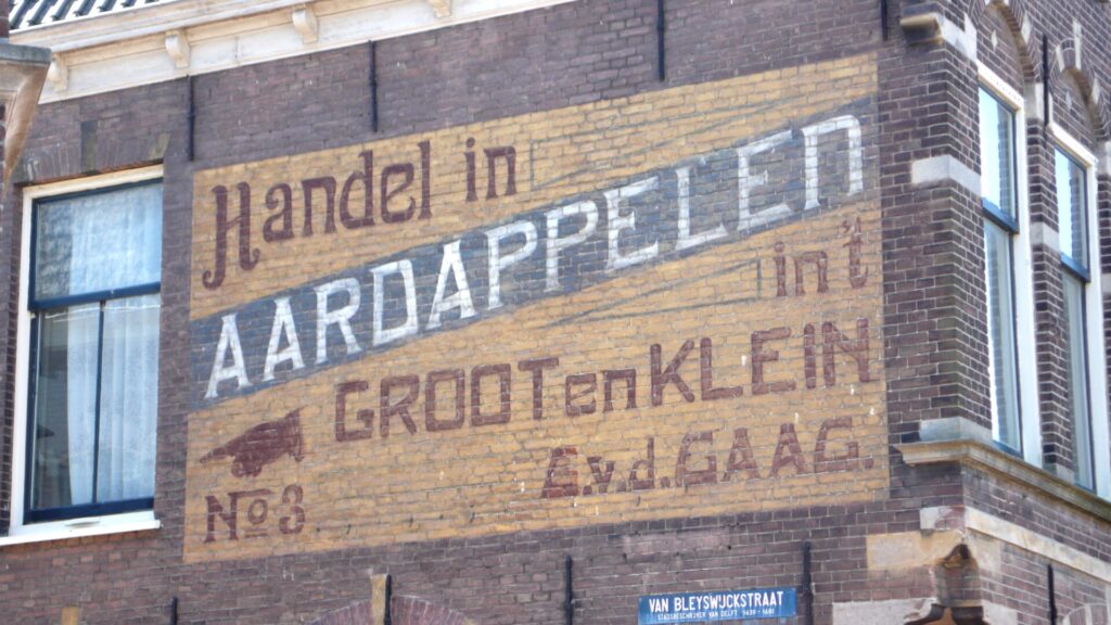 Fading painted sign on a brick building. It has a yellow background with most of the lettering in red set over it, and then a diagonal band of blue with some white lettering for the main word 'Aardappelen' in white. The the left of the sign is a manicule (pointing hand) illustration directing people to the street to the left of the sign.