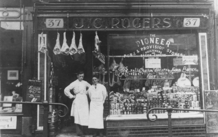 Archival photo of a butchers shop with meat hanging in the doorway where two butchers are posing for the photo. There is a carved and gilded sign above the shopfront that gives the building number (37) and the business name, J.C. Rogers. Gilded letters in the window then detail some of the products available.