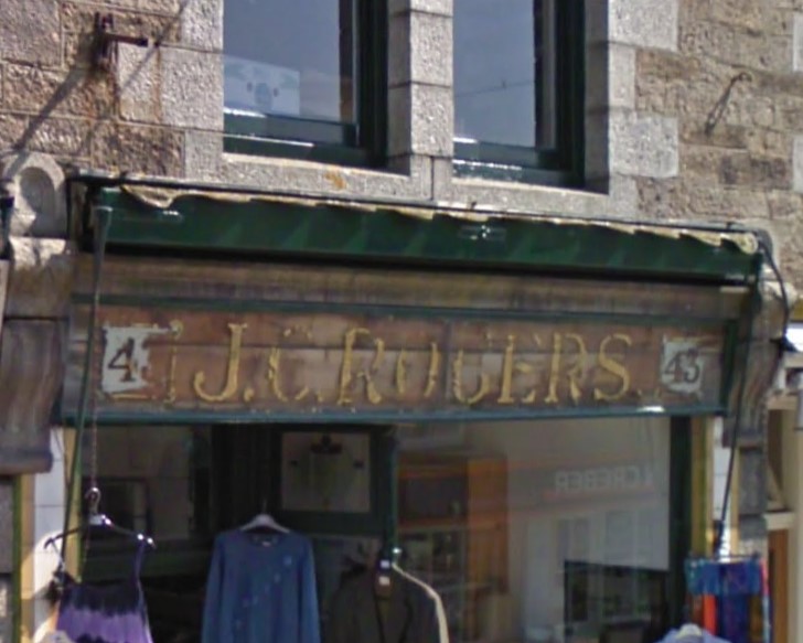 Slightly blurry screenshot from google streetview of a very decayed shopfront on which can be made out the door number (43) at either end and the business name, J.C. Rogers, in the middle. The letters appear to have been gilded in gold leaf on the reverse of a piece of glass before being installed.