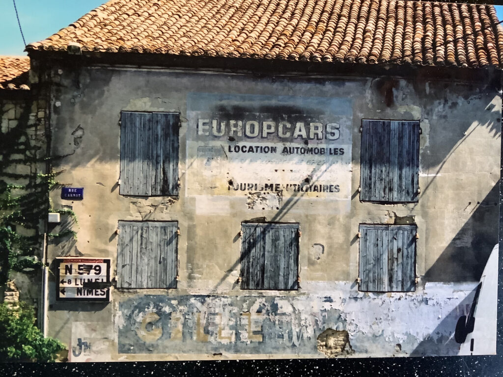 Run-down french house with shutters that also has the remains of fading painted signs on it. In addition to one that's barely legible along the bottom it one between the first floor windows that reads 'Europcars, Location Automobiles'.