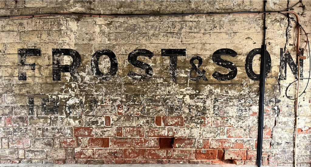 Brick wall in poor condition and with fading black letters in block lettering. Clearly visible at the largest size are "Frost & Son", while beneath, faded almost to illiegibility, is the word "Ironmongers".