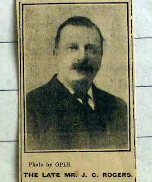 Black and white photo of a man from a newspaper clipping. He is wearing a suit and tie and has a moustache and short hair. Underneath the text says "Photo by Opie. The late Mr J.C. Rogers".