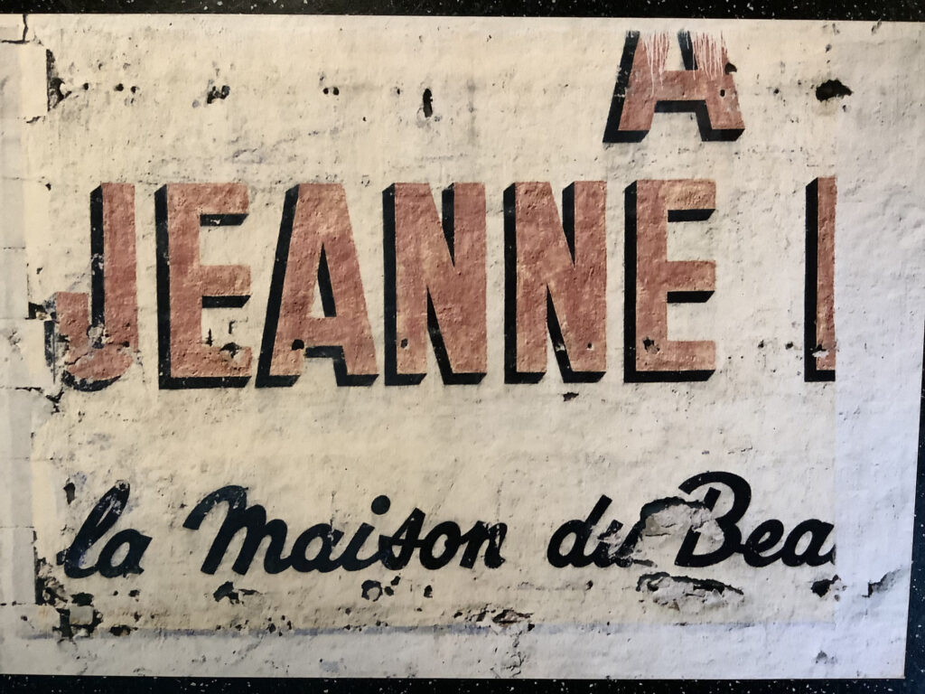 Detail of a fading painted sign on a decaying wall. The visible fragment has the letters 'A Jeanne' visible in red and, below that in black, 'la maison de Bea...'.