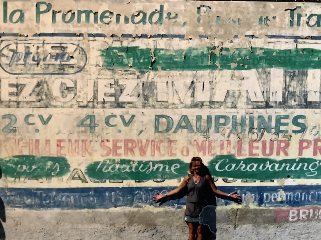 Woman standing in front of a tall wall that is covered in fading letters from different eras of signs that have been painted on it.