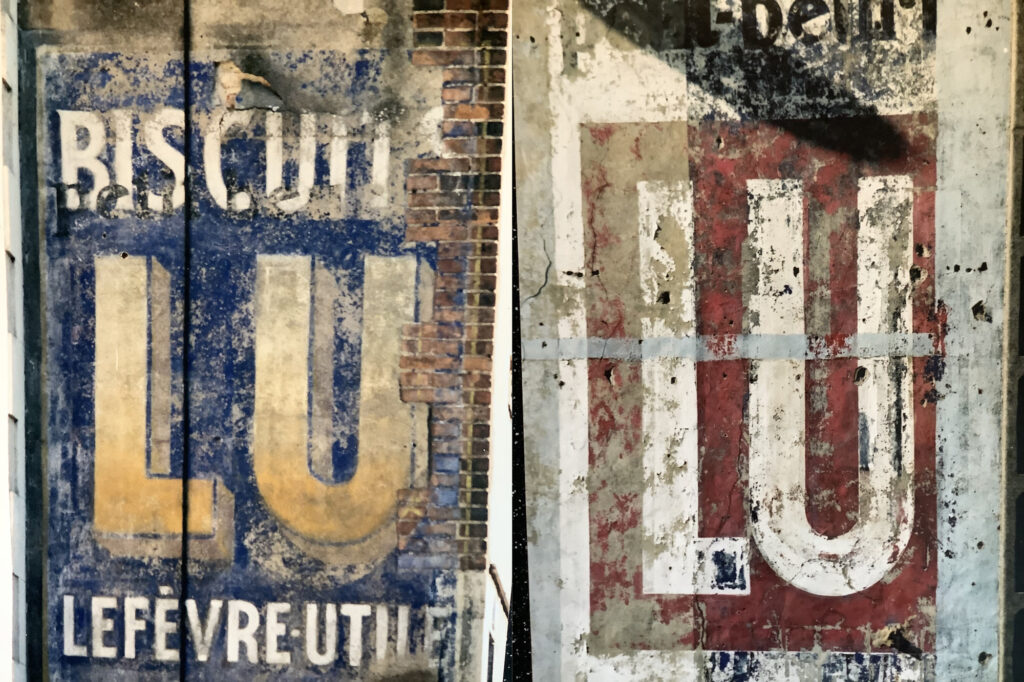 Close-ups of fading painted signs, each of which showing a different iteration of the Lu biscuits logo. The one on the left has the 'LU' letters in yellow on a blue background, while the one on the right has white letters on red.