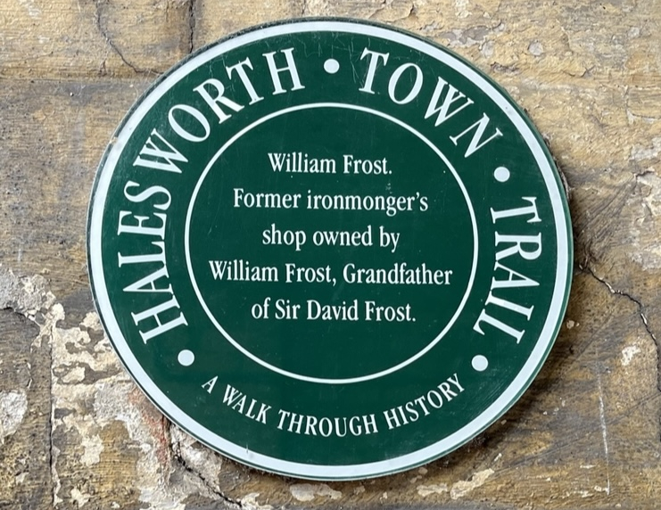 Circular green plaque with the "Halesworth Town Trail" arching over the top and "A Walk Through History" doing the same at the bottom. In the centre it says "William Frost. Former ironmonger's shop owned by William Frost, Grandfather of Sir David Frost".