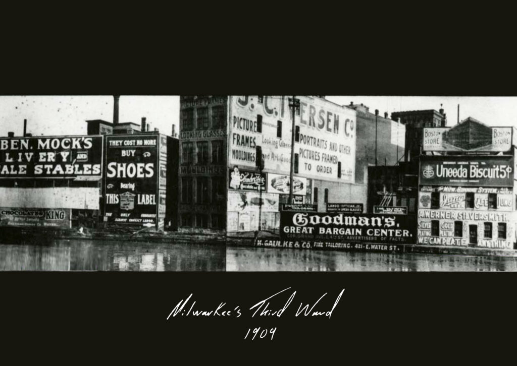 Black and white photo showing a riverside scene with numerous industrial buidlings almost completely covered with hand-painted signs for various different businesses including livery stables, biscuits, picture framers, a shoe manufacturer, and a silversmith.