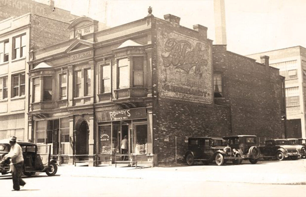 Black and white archival photo showing a corner building with a hand-painted wall sign for Blatz beer on it. Lined up in front of this are various vintage cars.