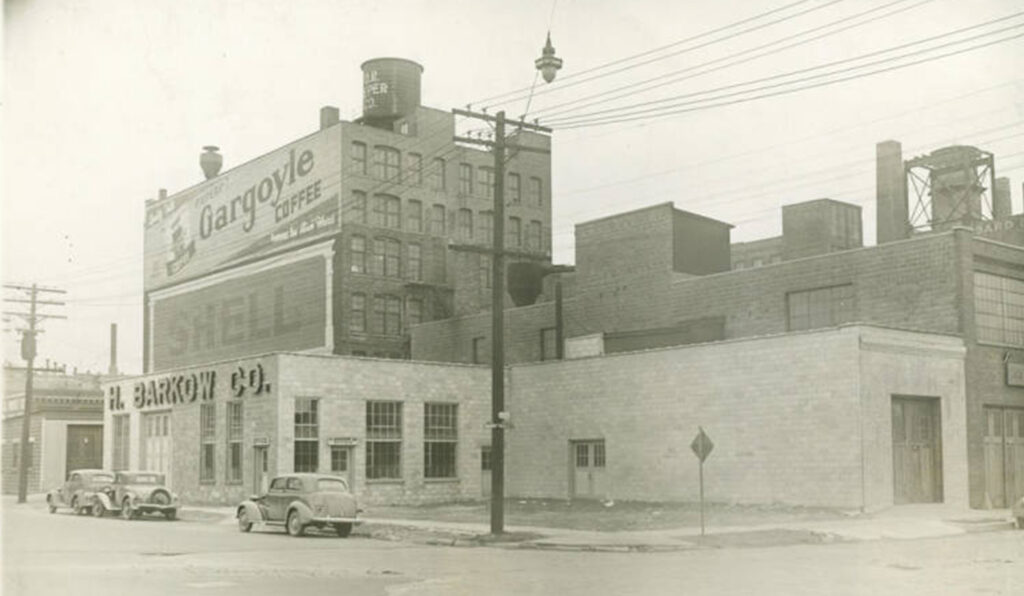 Archival photo showing large commercial buildings, one of which has two horizontal painted signs on the wall. One of these (top) advertises Gargoyle Coffee and the other is for Shell. There is then a protruding frontage below these with mounted channel letters for H. Barkow Co.