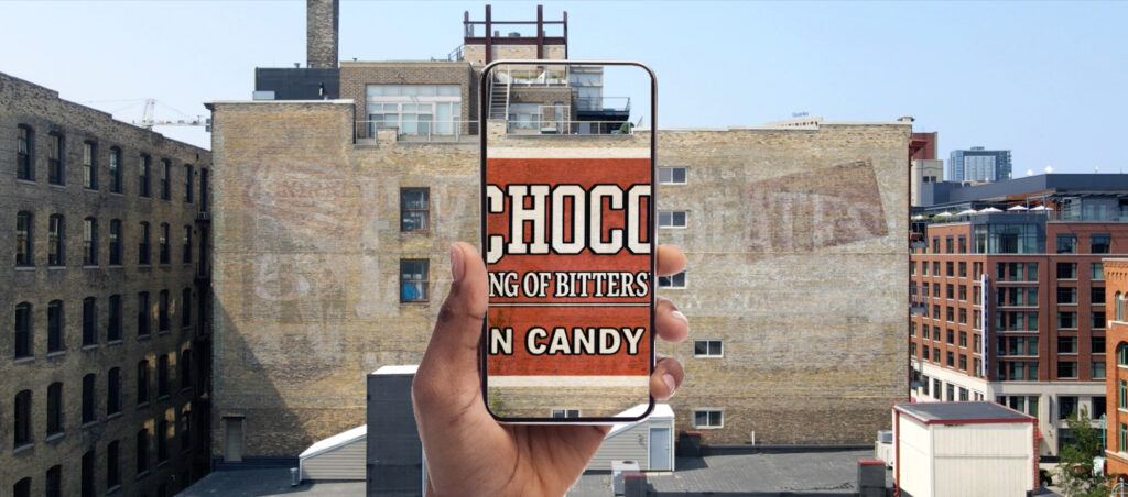 The same wall with a mobile phone being held in front of the middle portion. This shows the full-bodied artwork from the original Rex Chocolates sign to give an impression of how it would once have looked.